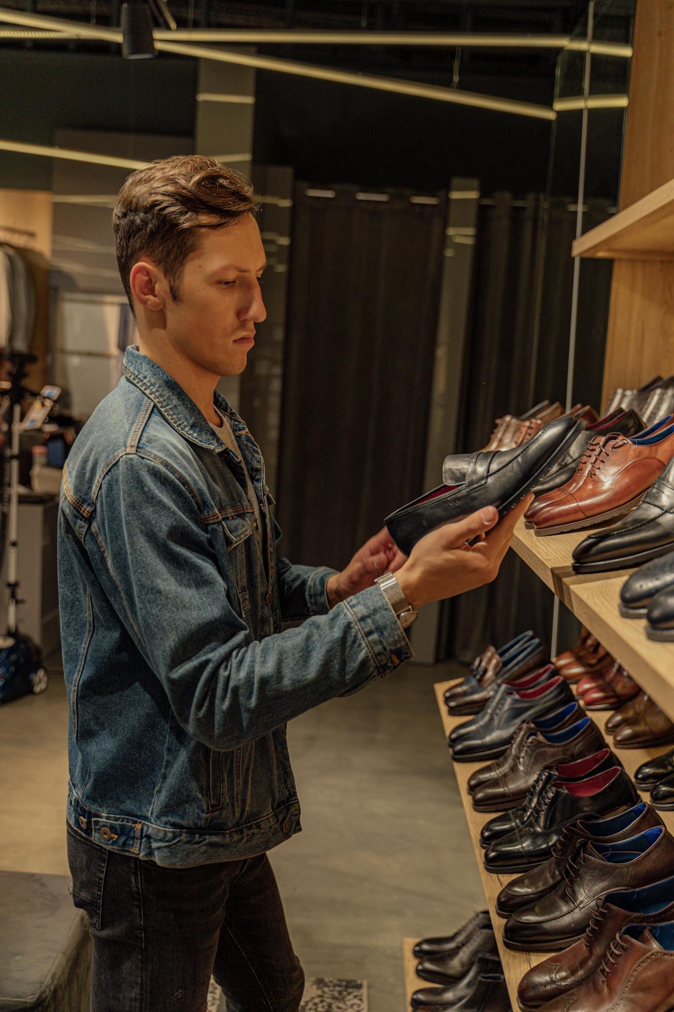 Young man in denim jacket browsing leather shoes in modern store.